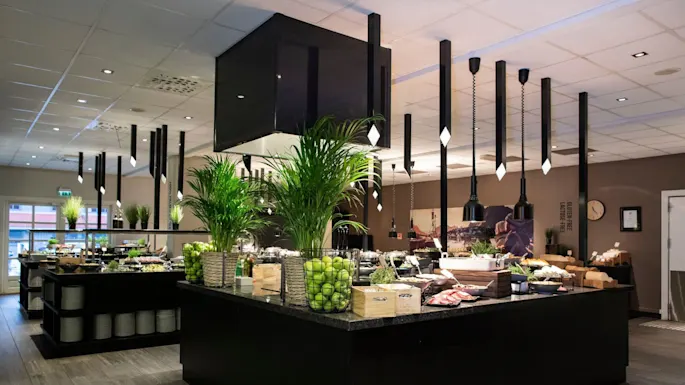 A spacious buffet area features counters laden with fresh greens, fruits, and various foods, all under stylish pendant lights. Green plants and baskets filled with apples add vibrant accents. Text on the wall reads: “GLUTEN-FREE LACTOSE-FREE”.
