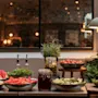 A vibrant buffet at Home Hotel Grand Bodø, featuring fresh fruits, vegetables, and pickled items, illuminated by a stylish lamp.
