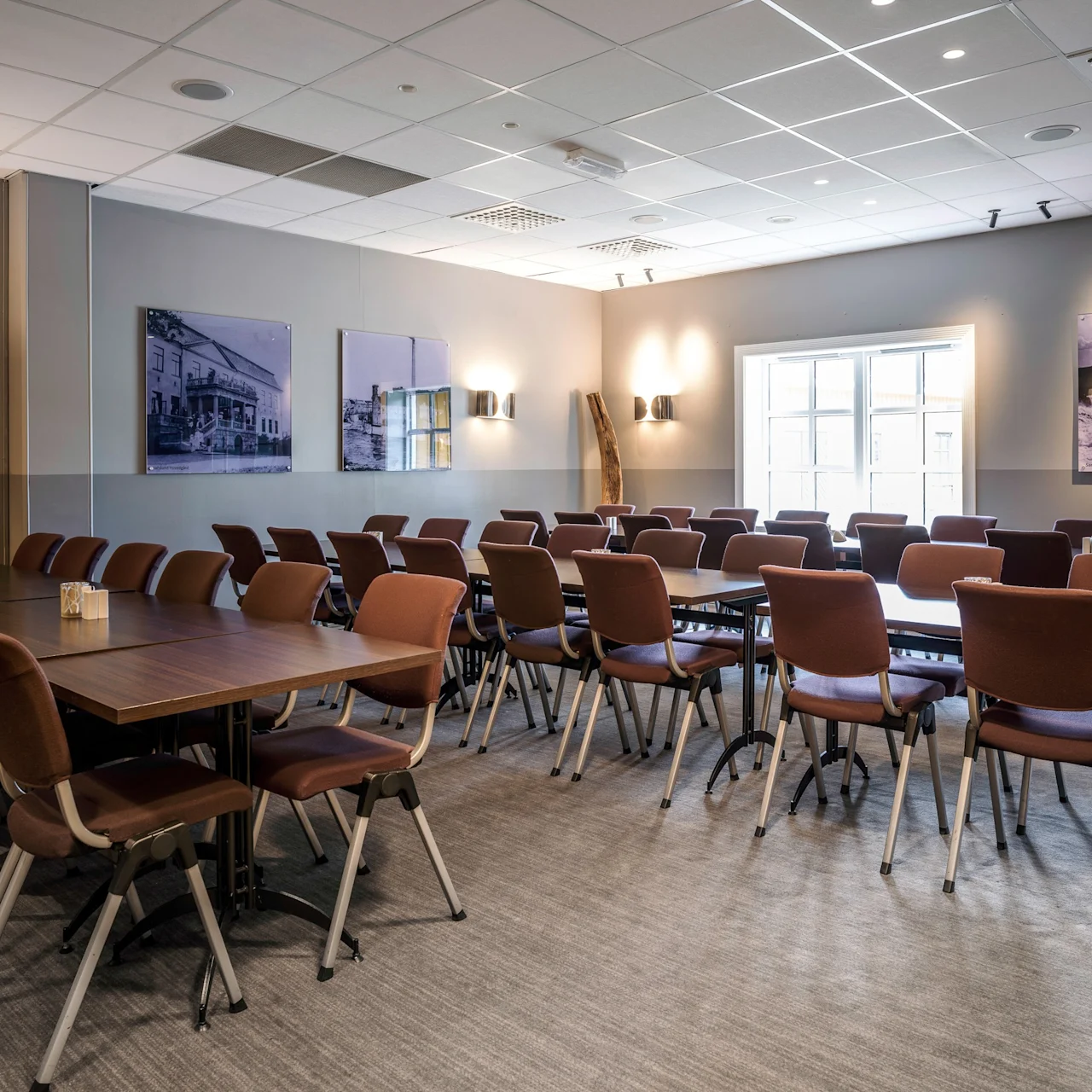 Brown chairs face a projector screen in a conference room, arranged in rows with a wooden floor and photographs on walls, lit by ceiling lights and natural window light.