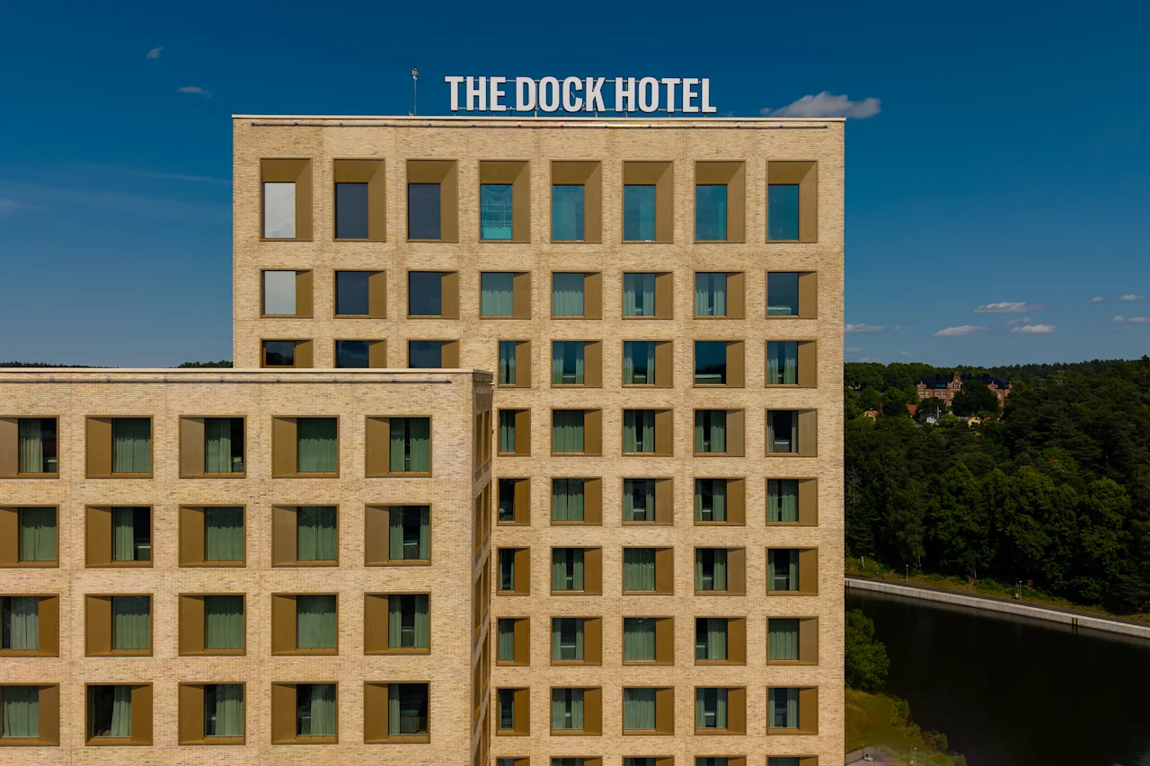 The Dock Hotel, en moderne bygning med mange vinduer, står mot en blå himmel med en grønn skogkledd bakgrunn.
