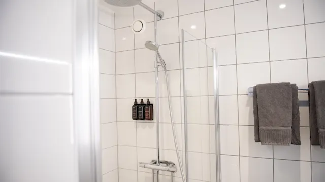 A showerhead with a sliding bar is mounted on a tiled bathroom wall. Three bottles sit on a shelf, and two gray towels hang on a nearby rack.