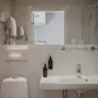 Handfat med kran i ett vitkaklat badrum; flankerad av tvåltvålpumpar och glas. I närheten finns en toalett, väggmonterad hårtork och handdukshängare som förstärker hotellets enkla, funktionella estetik.