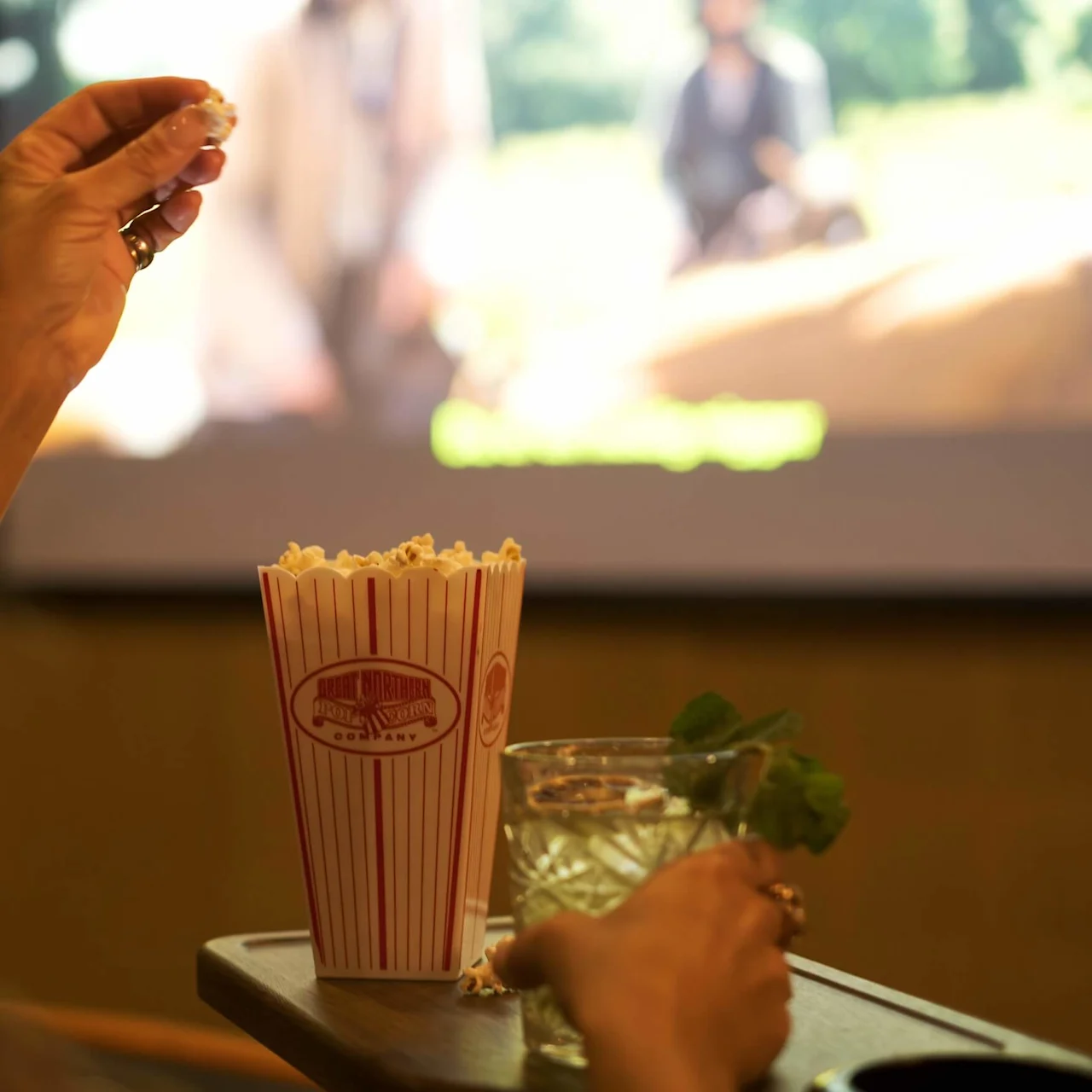 Popcorn is being eaten by a person sitting in front of a screen showing a movie. A striped popcorn container and a drink with mint garnish are on a tray.