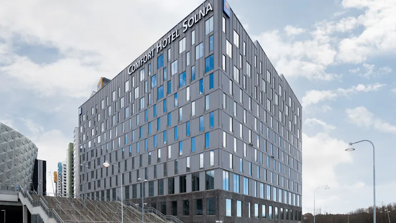 A tall, modern building labeled "COMFORT HOTEL SOLNA" stands prominently on a city street, with a cloudy sky above and adjacent urban structures.