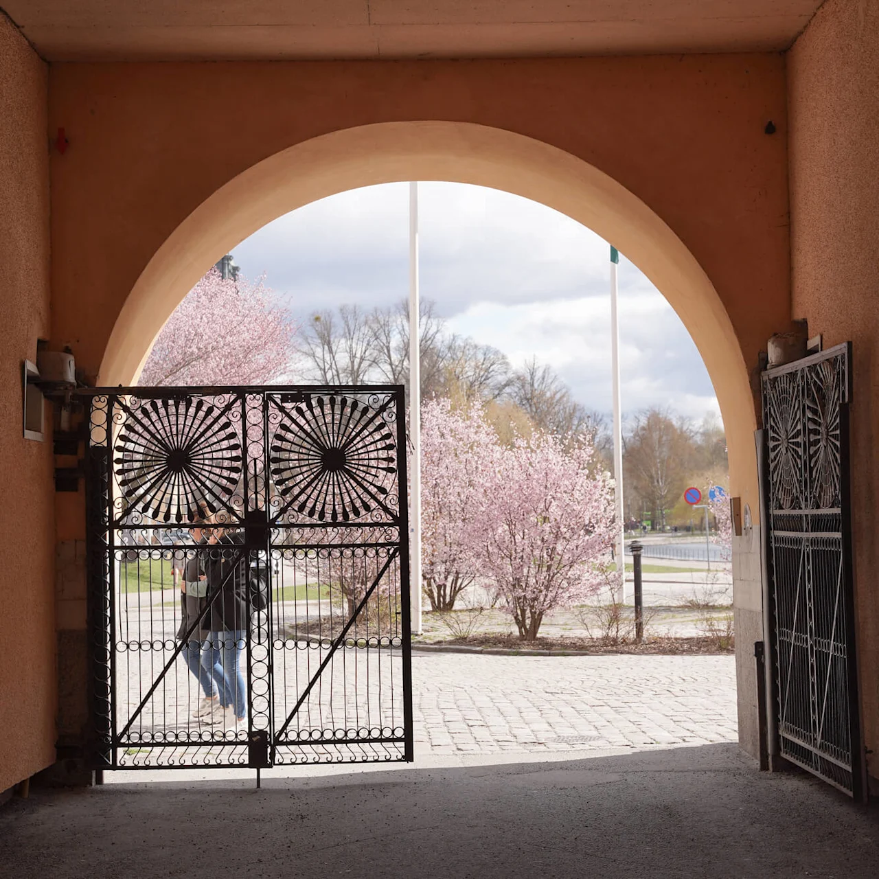 Person passerar genom en utsmyckad grind, sett från en tunneliknande valvöppning som leder till en ljus, blommande utomhusscen.