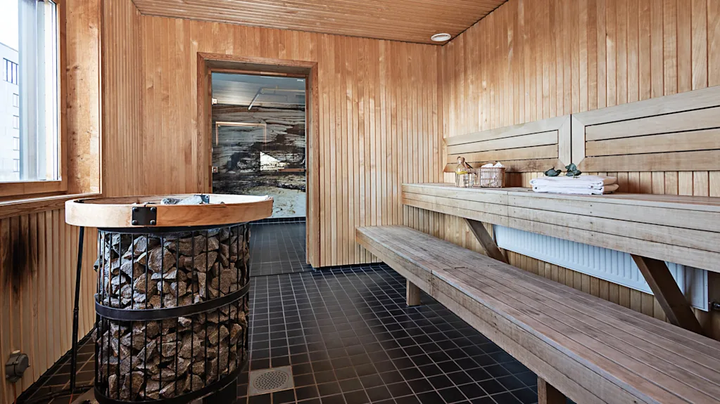 A wooden sauna with a central stove heating stones, and wooden bench seating along the sides. The sauna is well-lit through a window, with folded towels on the bench.