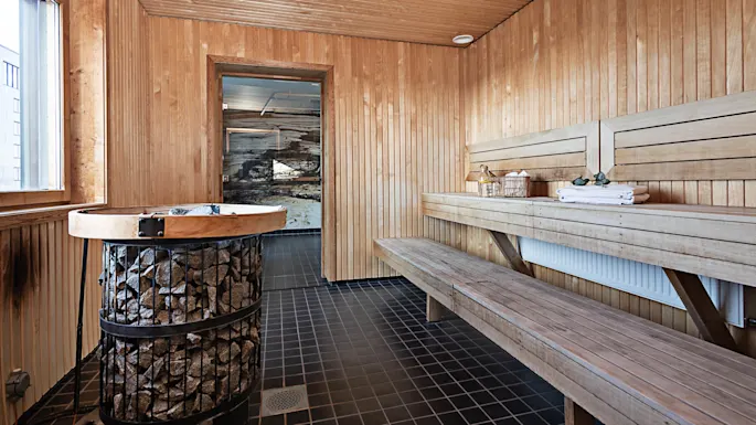 En trebadstue med en sentral ovn som varmer steiner og trebenker langs sidene. Badstuen er godt belyst gjennom et vindu, med sammenfoldede håndklær på benken.