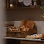 Ett trälåda fylld med blandat bröd står på en köksbänk. Burkar med apelsin och röd sylt står bredvid, under en hylla med dekorativa växter.