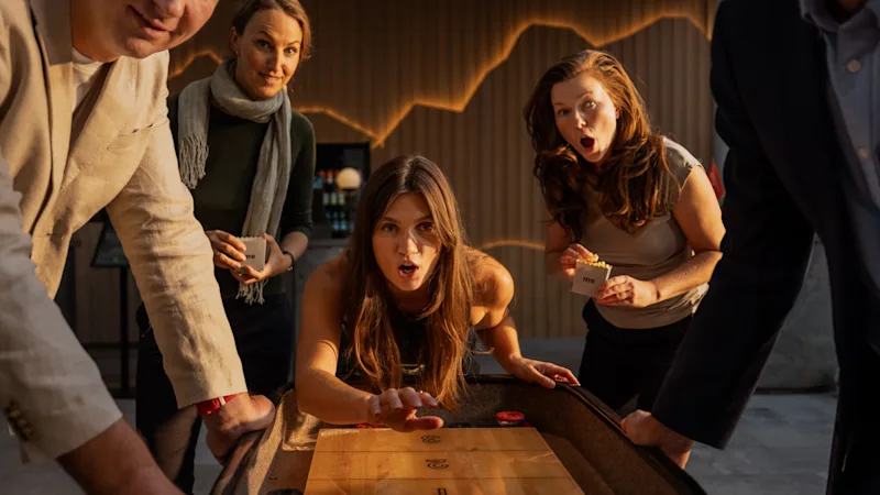 En grupp på fem personer samlade runt ett shuffleboard-bord, en person är på väg att göra ett drag. Andra tittar intensivt, håller drycker i ett varmt belyst rum.