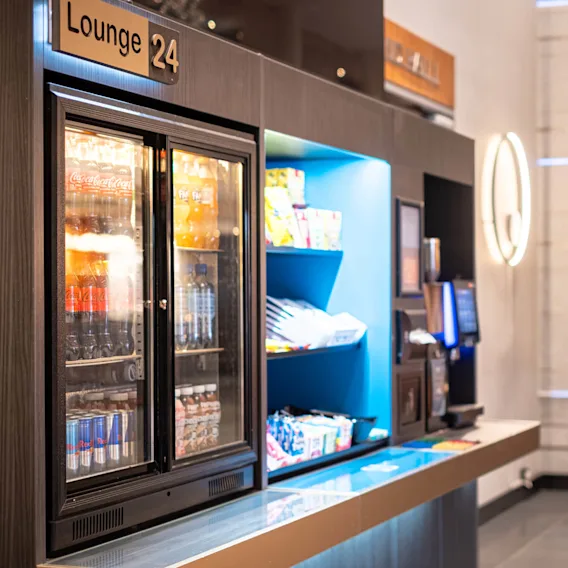 Kjøleskap med drikkevarer ved siden av hyller med snacks; konteksten er et moderne loungeområde merket "Lounge 24."