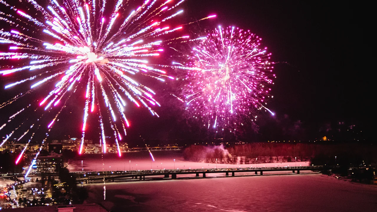 Fireworks explode in vibrant red and white over a city at night, illuminating a bridge and river below, creating a festive atmosphere.