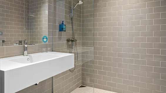 Sleek white sink with a metallic faucet sits in a tiled bathroom. Nearby, a walk-in shower has a glass partition. Wall-mounted pump bottle labeled: "DOVE MEN+CARE".