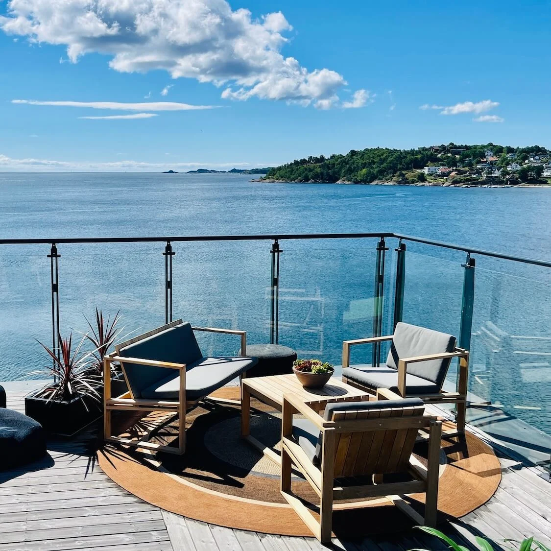 En terrasse med moderne møbler har udsigt over et glitrende hav under en klar blå himmel.