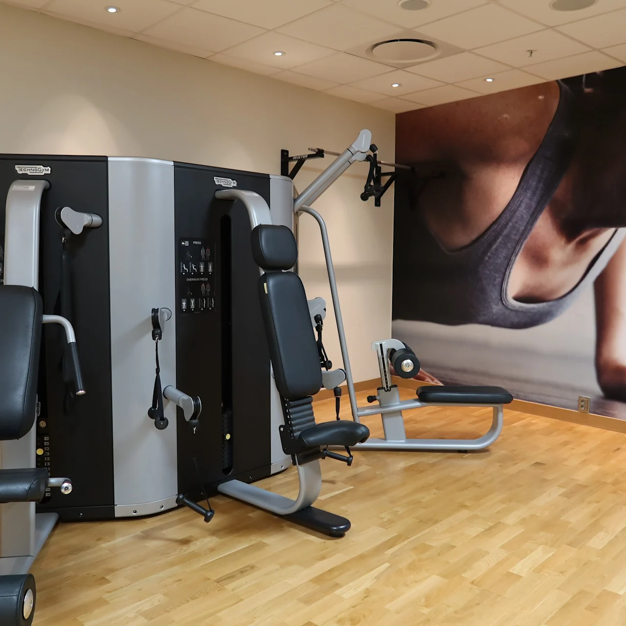 A multi-station gym machine with various exercise attachments sits on a wooden floor. Adjacent is a large wall poster showing a close-up of a person in a push-up position.