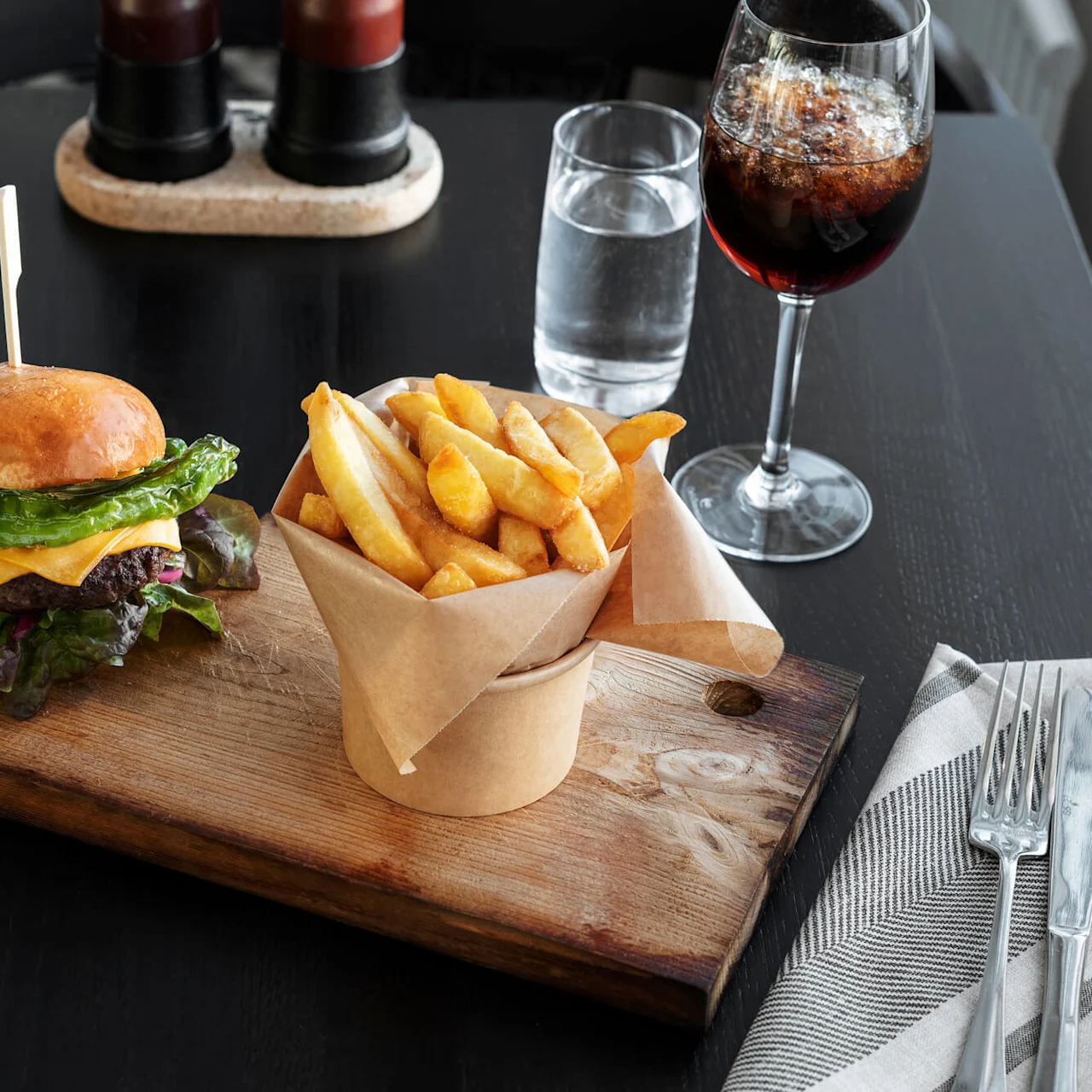 Burger med salat og ost på en træbræt, ledsaget af pommes frites i en papirkop. Et glas cola, et glas vand og bestik på en serviet på et mørkt bord.