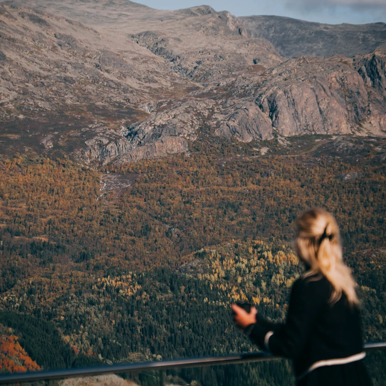 A person with blonde hair observes a vast mountain landscape covered in autumn foliage, standing behind a railing. Clouds hover above the rocky peaks.