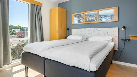 A neat bedroom with a double bed, wooden furniture, and framed pictures on a blue wall, bright day outside.
