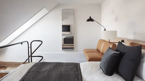 Bed with dark cushions sits in a minimalist room; a couch and wall lamp are nearby, facing a wall mirror reflecting a TV. A sloped ceiling with a skylight is above.