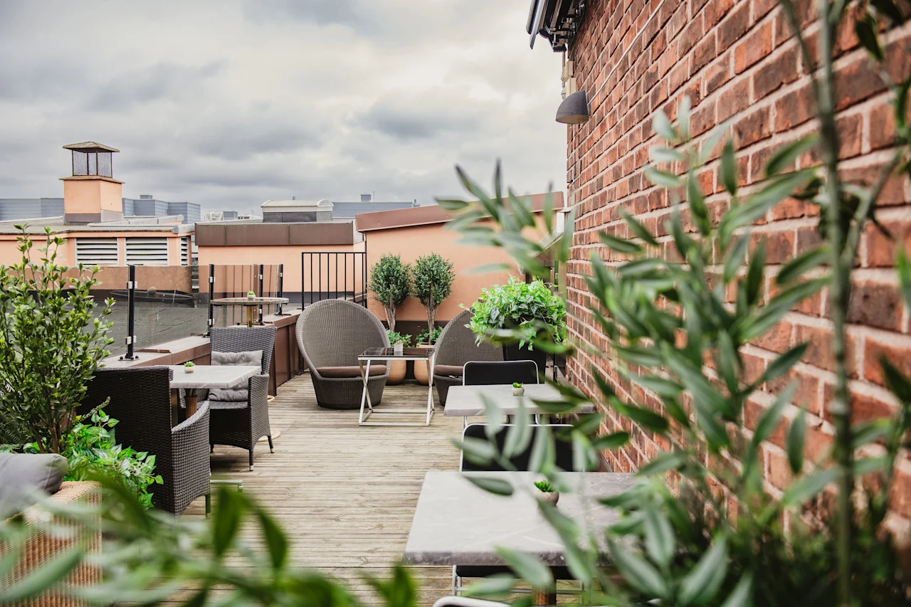 A rooftop terrace with outdoor seating, lush green plants, and a brick wall under a cloudy sky.