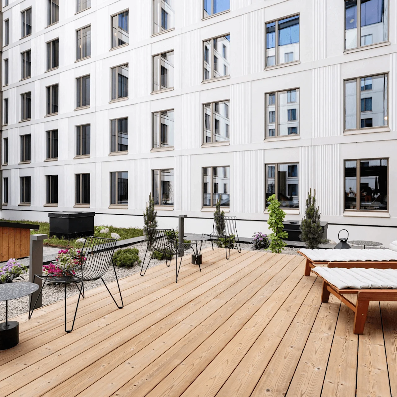 Sundeck features lounge chairs and tables on a wooden terrace amid a modern courtyard with white multi-story building and greenery.