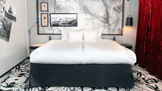 A neatly made double bed with white linens, framed pictures on the wall, a red velvet curtain, and a patterned carpet in a bedroom.