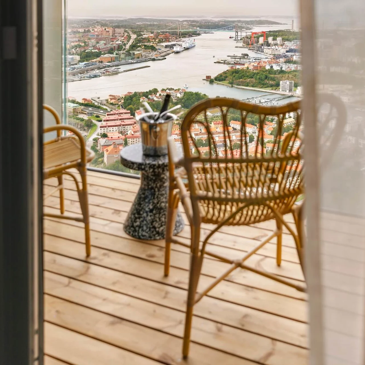 Wickerstolar och ett bord med en champagnehink står på en träbalkong. Utsikten visar en flod, stadslandskap och avlägsna kullar under en delvis molnig himmel.