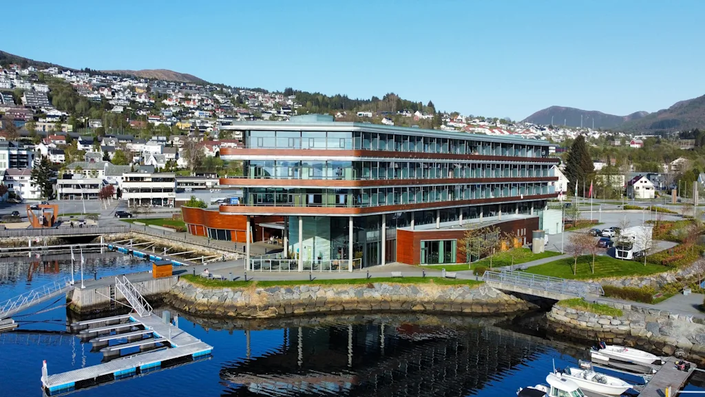 En modern, flerfaldig byggnad med stora glasfönster står vid en marina med flytbryggor. Bostadshus och gröna kullar syns i bakgrunden.