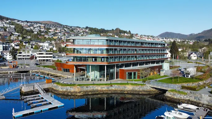 A modern, multi-story building with large glass windows stands by a marina with floating docks. Residential houses and green hills are visible in the background.