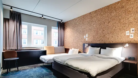 A neatly made bed is in a modern hotel room, accompanied by a small sofa and chair. Natural light enters through large windows, complemented by a textured cork wall with small art pieces.