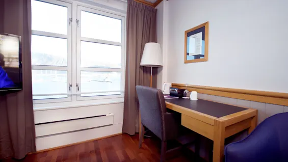 Et hotelværelses interiør med et træskrivebord, stole og et vindue der afslører et bjerglandskab og vand.