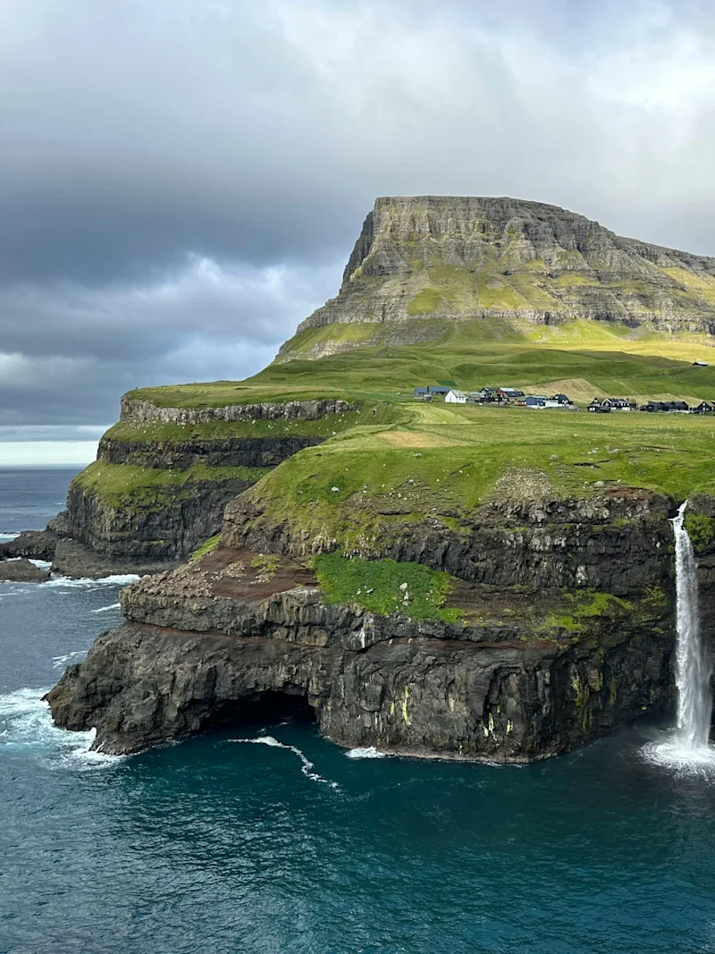 En fantastisk udsigt over et vandfald, der kaskader fra en grøn klippe ned i det mørkeblå hav under en overskyet himmel. Små huse er synlige på klippetoppen.