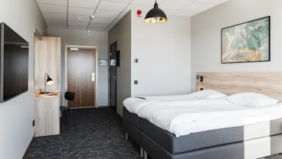 En säng med vita lakan står i ett modernt hotellrum med en väggmonterad TV, ett skrivbord med lampa och abstrakt konst. Grå matta och varm belysning skapar en lugn atmosfär.