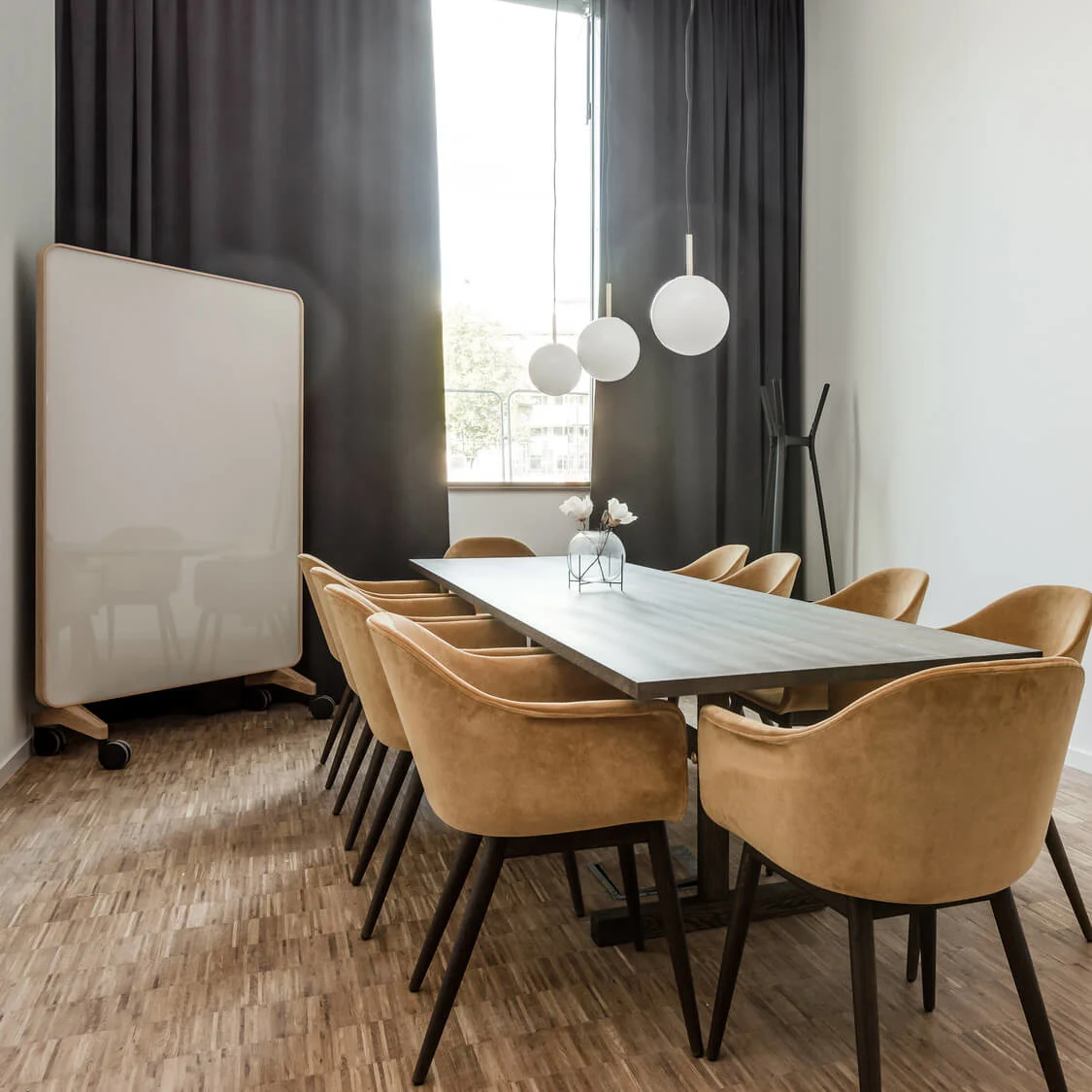Et langt konferancebord med beige stole omkring, under tre hængende sfæriske lamper i et moderne møderum med trægulv og et stort vindue med sorte gardiner.