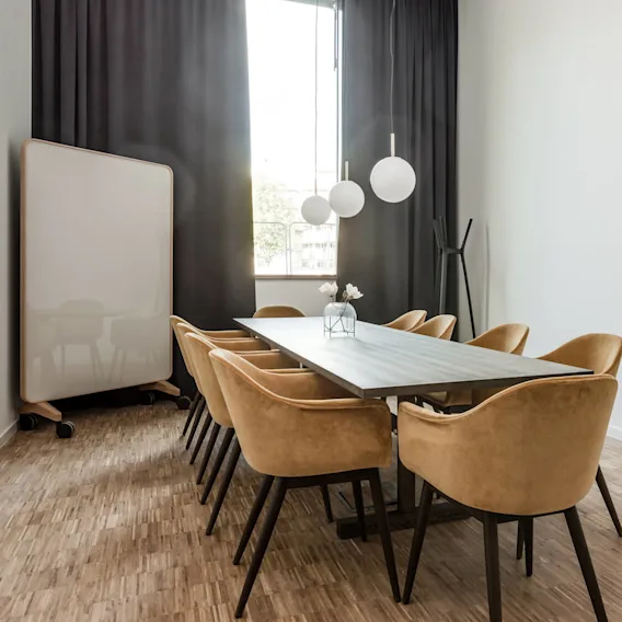 A long conference table with beige chairs surrounds it, under three hanging spherical lights in a modern meeting room with wooden flooring and a large window draped in black curtains.