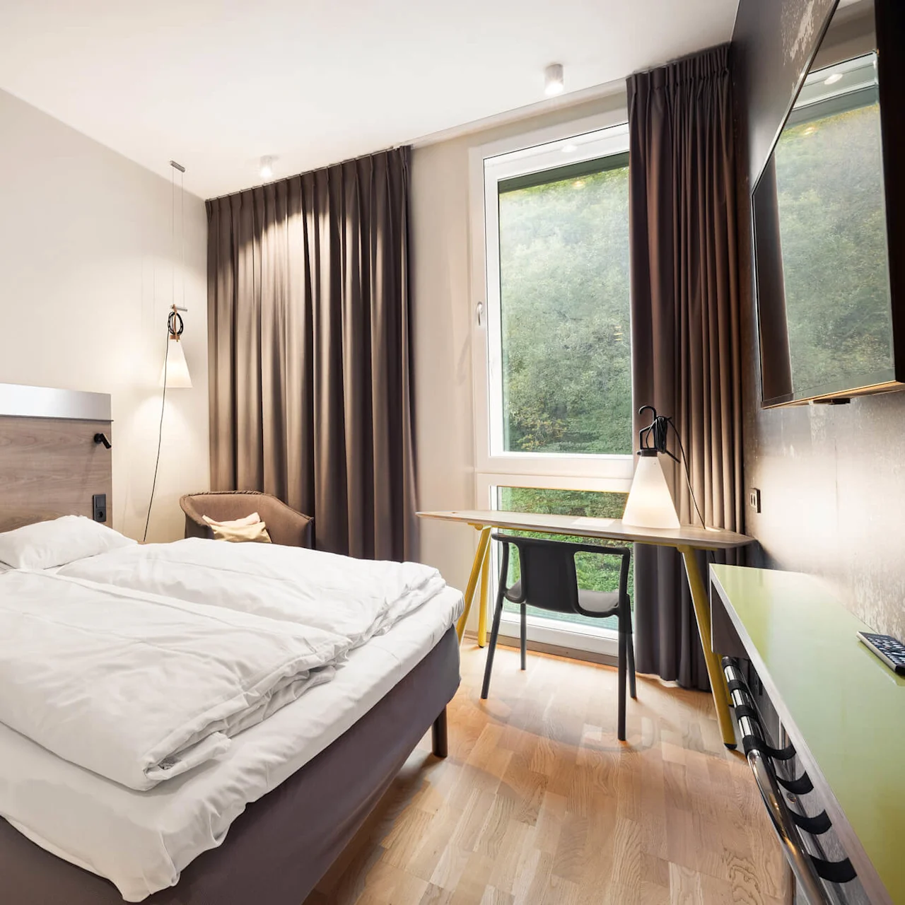 A neatly made bed stands in a hotel room, accented by a large window with brown curtains. A small desk and chair sit nearby, with a lamp providing ambient lighting.