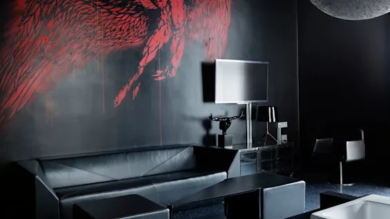 Black leather sofa sits against a dark wall with red winged figure art. A TV is mounted nearby, with a modern lamp and chair completing the sleek living room setting. 