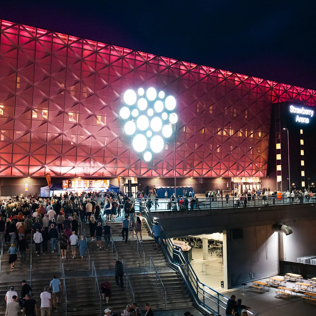 Suuri ihmisjoukko kävelee portaita ylös kohti valaistua areenaa yöllä. Areena.