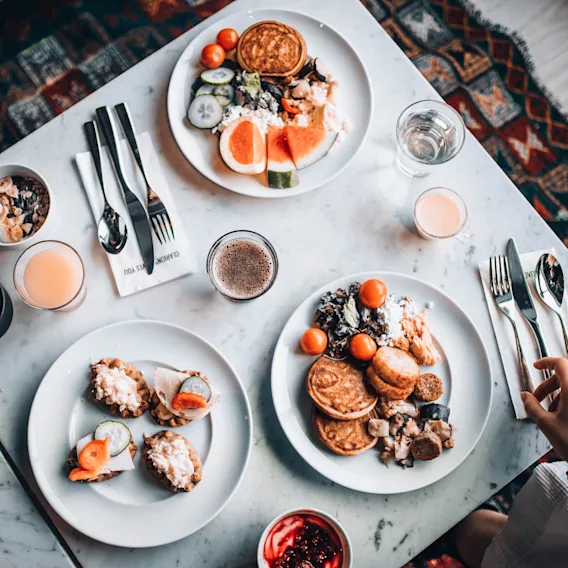 Tre tallerkener med frokostmat, inkludert pannekaker, frukt og yoghurt, står på et marmorbord med bestikk. Drikker inkluderer kaffe og juice. En hånd strekker seg etter en gaffel.
