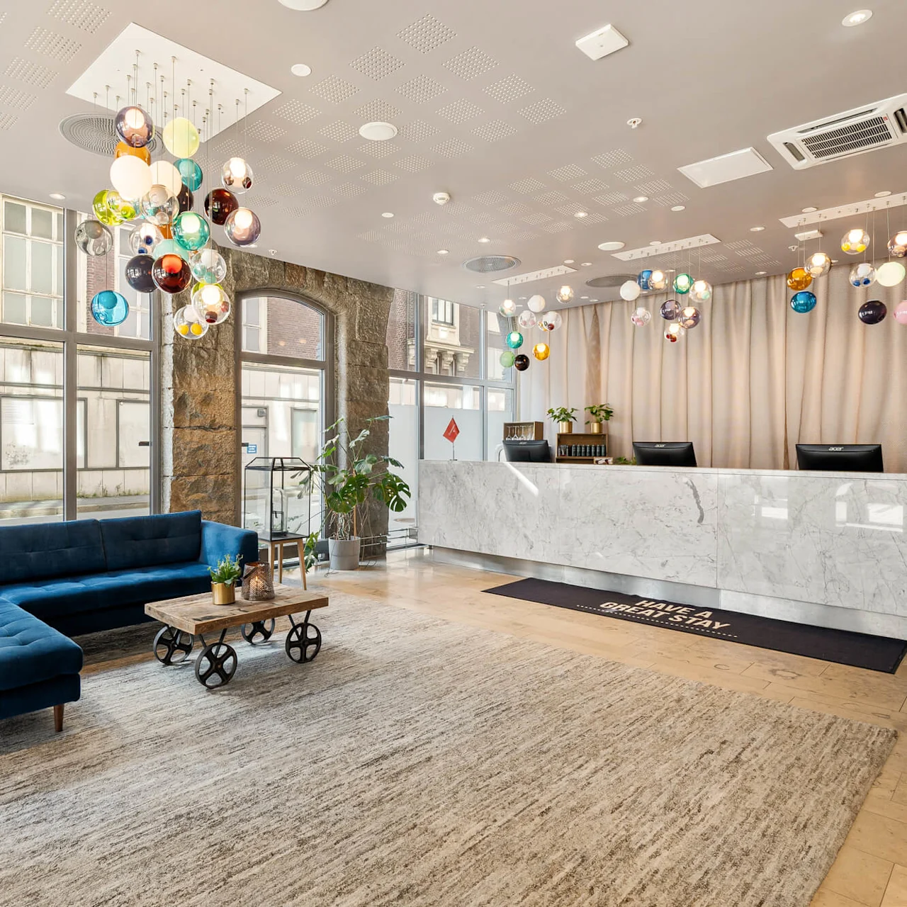 En marmorreception med farverige pendellamper ovenpå står i et moderne lobbyområde med en blå hjørnesofa og et træbord. "HAVE A GREAT STAY" står på gulvmåtten.