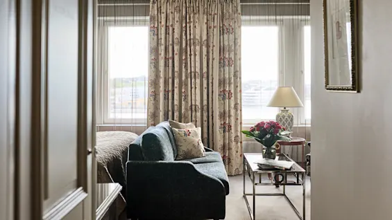 Et vindue med blomstrede gardiner bader et hyggeligt rum i lys, med en polstret sofa, glasbord med blomster og sengelampe, hvilket skaber en rolig atmosfære.