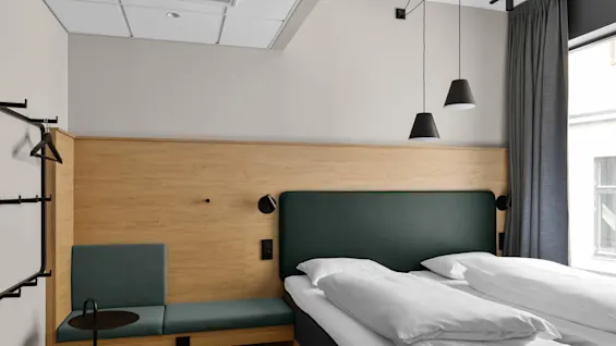 Ett modernt hotellrum med en träpanel har en dubbelsäng med vita sängkläder, en liten stoppad bänk, pendellampor och ett fönster med grå gardiner.