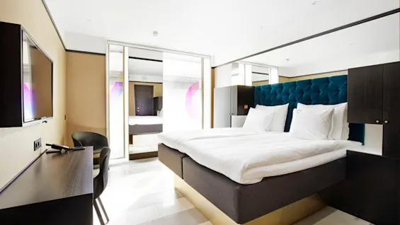 En dobbeltseng med mørkt sengegærde står centreret i et moderne hotelværelse. Til venstre er der et skrivebord og en stol med et stort fladskærms-TV på væggen.