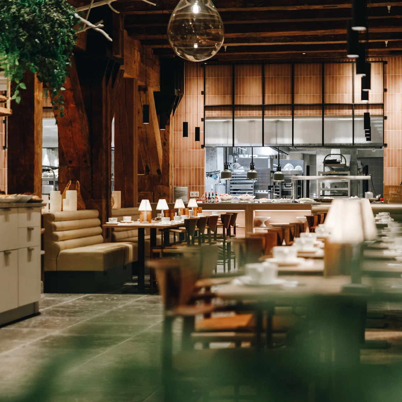 A spacious, modern restaurant interior features empty wooden tables and chairs, with a central open kitchen visible. Hanging plants and soft lighting create a warm atmosphere.