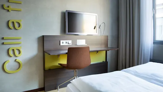 Television mounted on a wall, above a wooden desk with a brown chair in a modern hotel room. "Call a friend" is written vertically on the left wall. 
