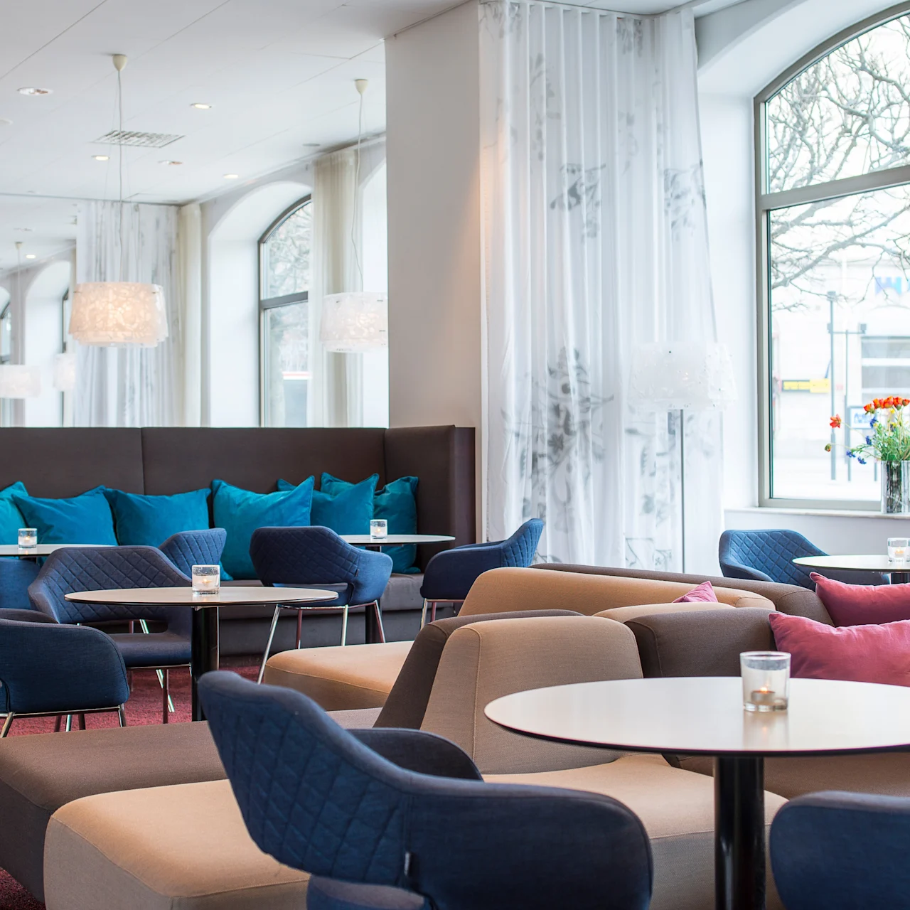 Komfortable lounge-stoler omgir små runde bord i et lyst rom med store vinduer, blå og rosa puter og myk belysning. Blomster er synlige på et bord.
