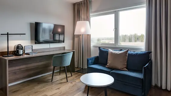 A cozy room features a blue sofa with a brown cushion and a round coffee table, positioned next to a window showing a landscape view. Nearby is a desk with a TV, coffee machine, and a chair.