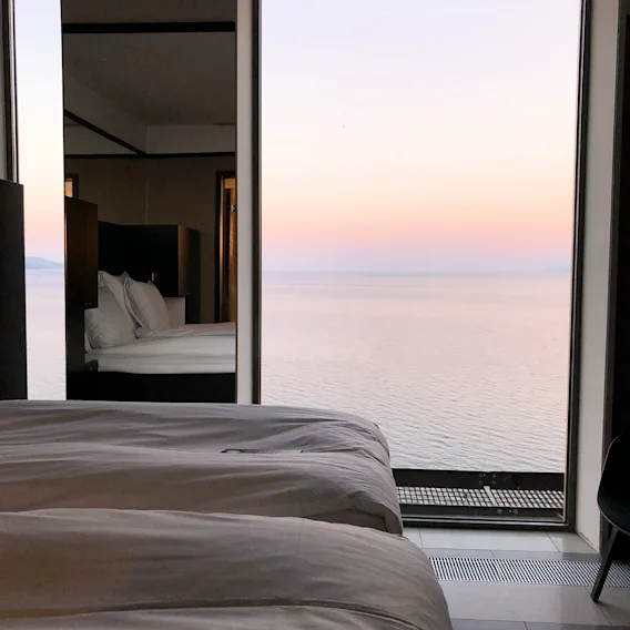 En mysig hotellsäng med en fantastisk utsikt över ett lugnt hav under en mjuk rosa och orange solnedgång.