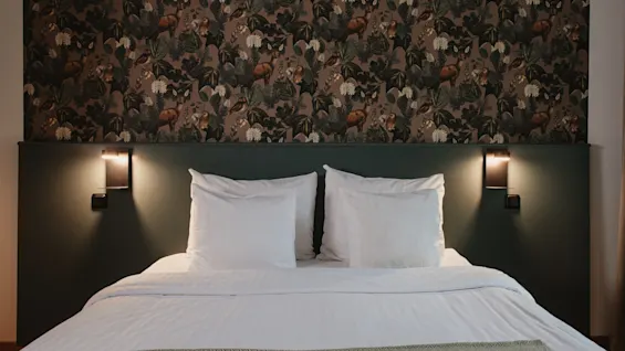 A cozy hotel room at Home Hotel Uppsala with a bed, white linens, and a nature-themed wallpaper. Wall lights provide a warm glow.