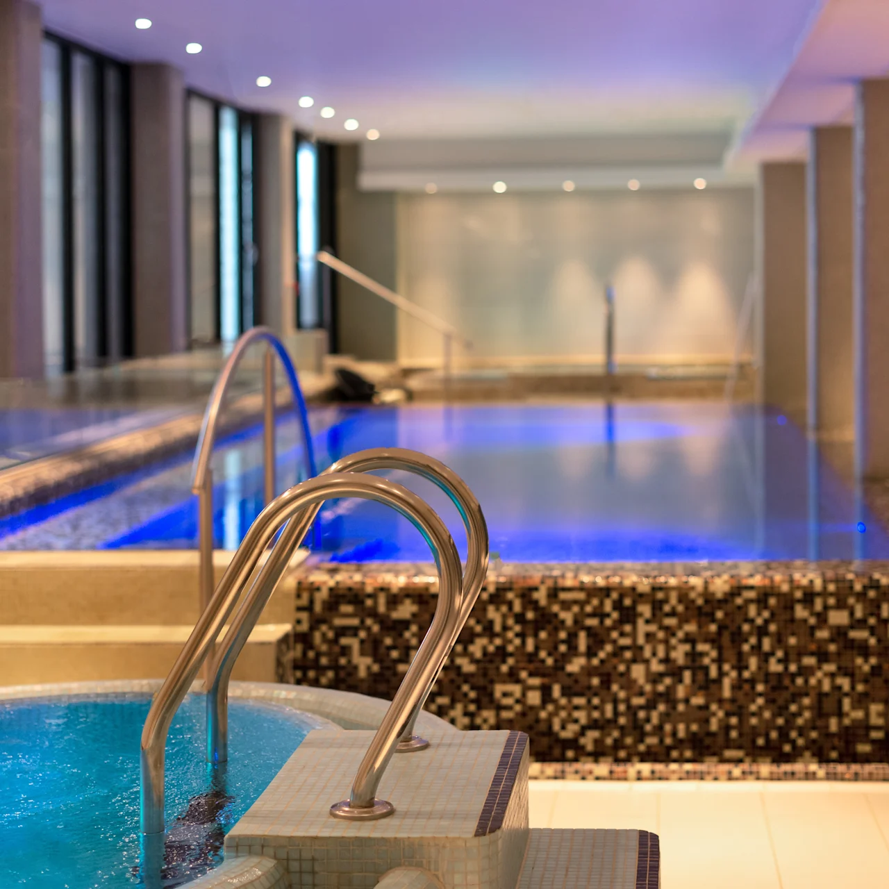 Hot tub with bubbling water, next to a large, illuminated indoor pool. Surrounded by tiled flooring and windows with soft overhead lighting. Quiet, serene spa environment.