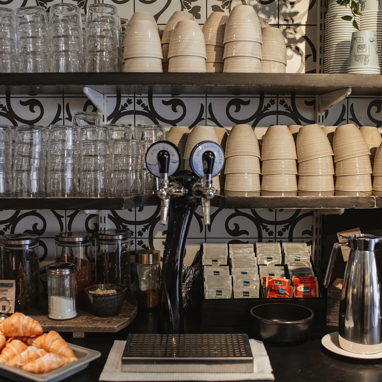 Café counter with stacked cups and glasses on shelves, and two coffee dispensers below. Croissants on a tray and various condiments are arranged beside a thermos, against a decorative wall.
